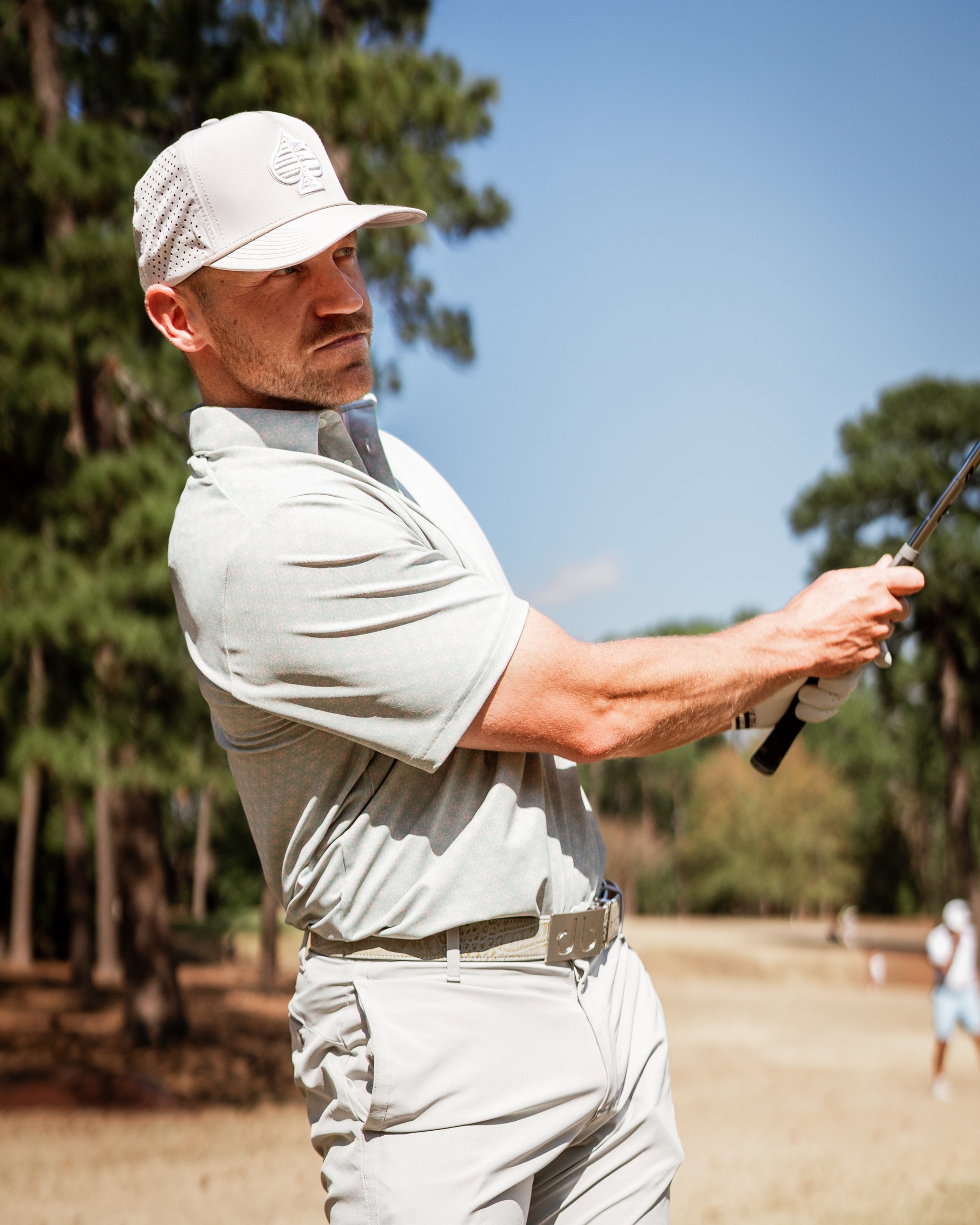 A golfer swings a club on the course, wearing a light gray polo and a matching cap, showcasing a stylish golf look.