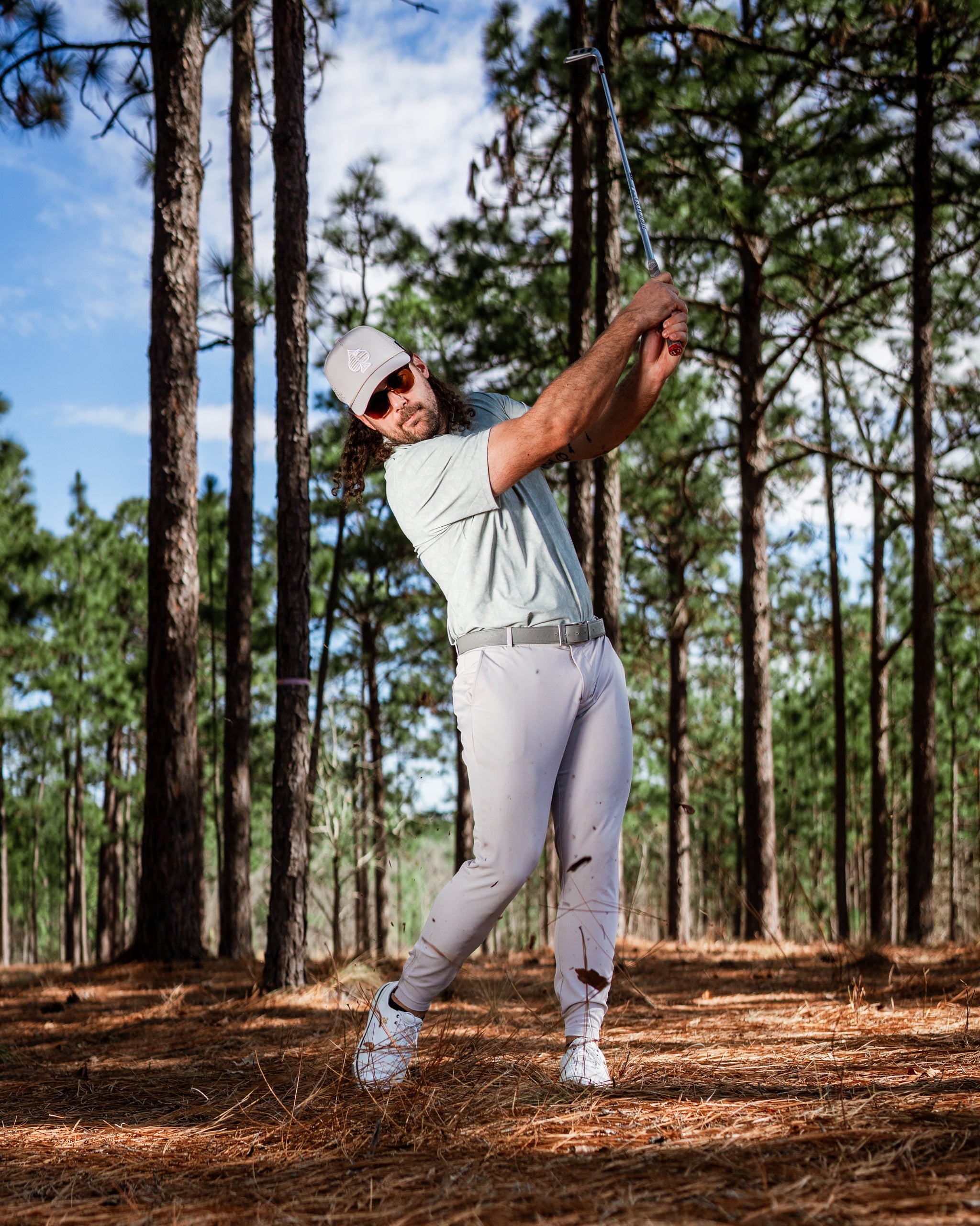 Golfer swinging a club in a pine forest, wearing a light green shirt and white pants, showcasing a stylish golf look.