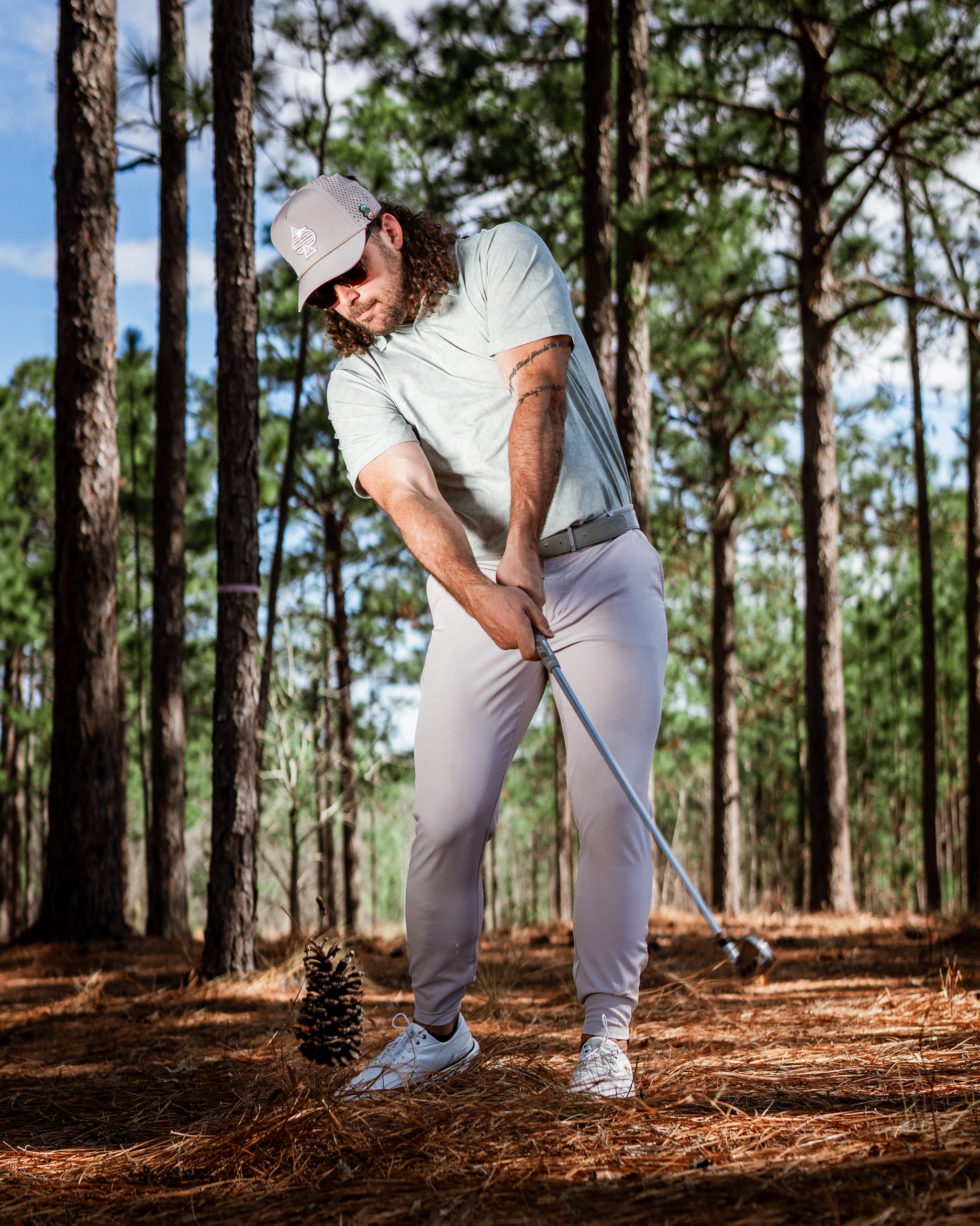 Golfer in a light green shirt and cap swings a club at a pinecone in a forest setting, surrounded by tall trees.