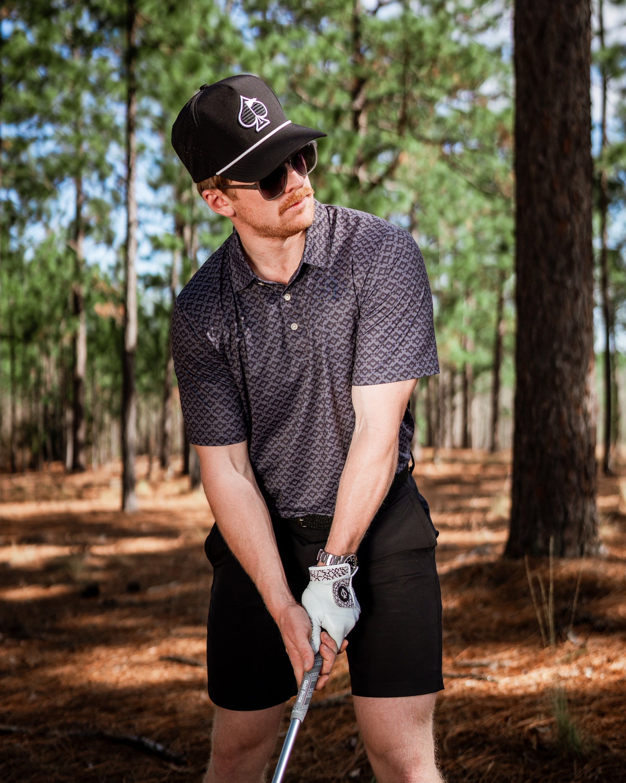 A golfer in a slate mosaic polo shirt and black shorts prepares to swing in a wooded area, showcasing bold golf style.