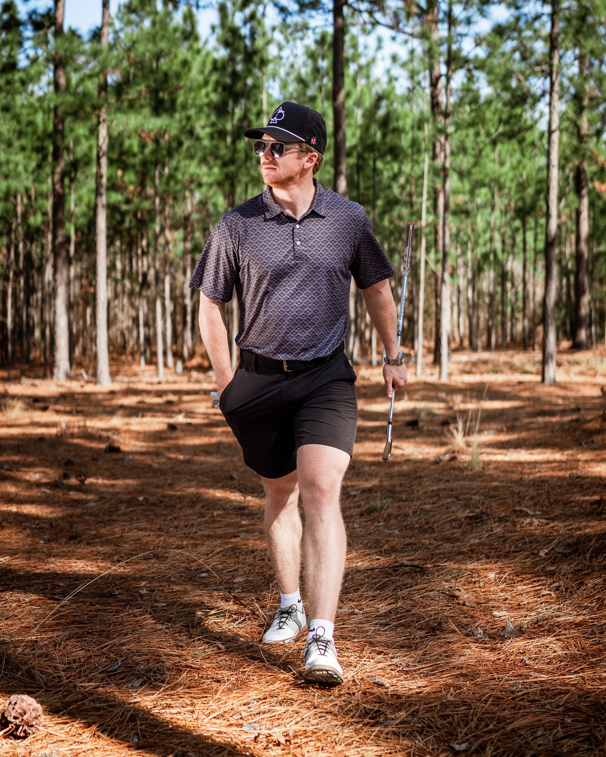 A golfer wearing a slate mosaic polo and black shorts walks confidently through a pine forest, holding a golf club.