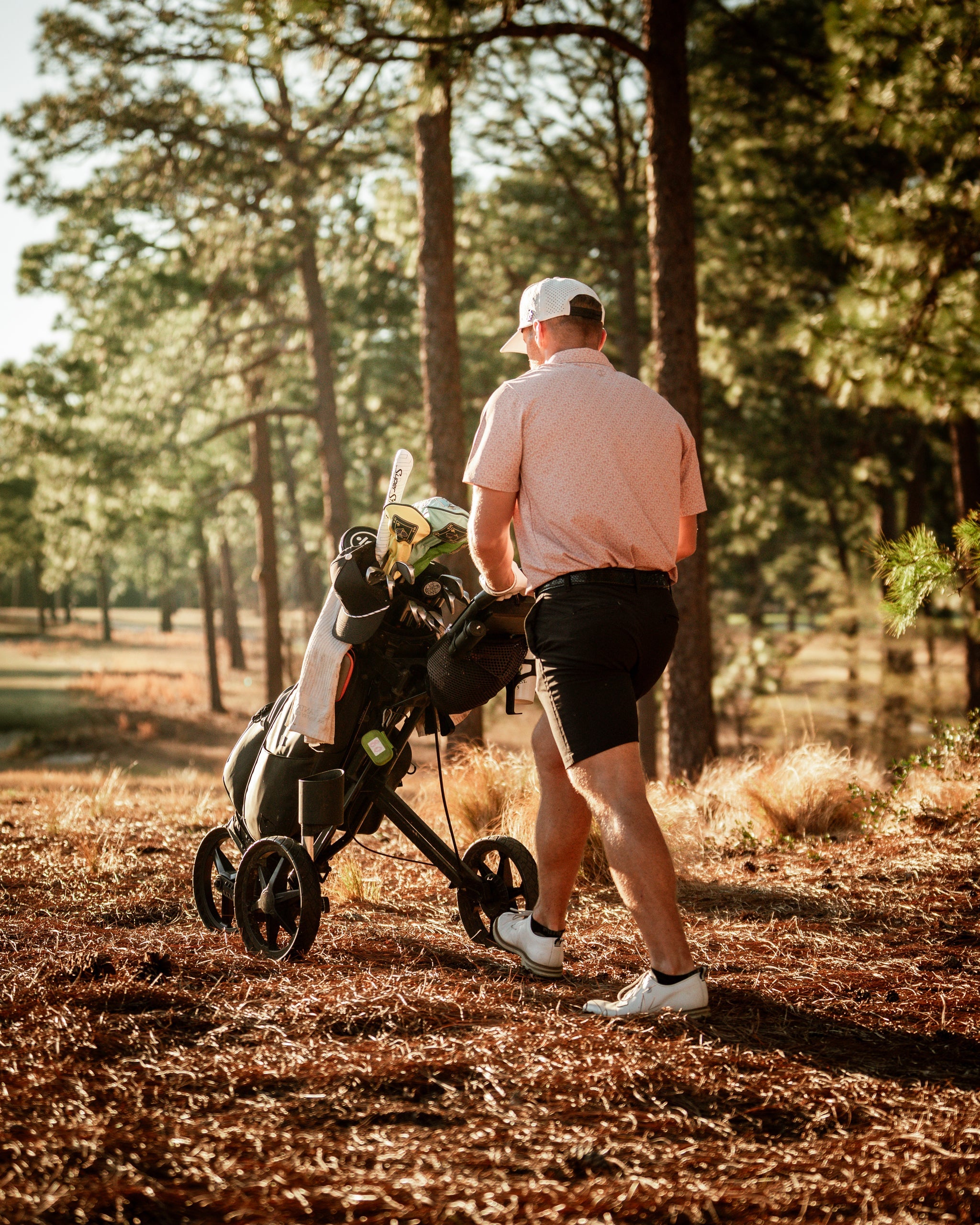 A golfer walks with a golf cart in a sunlit forest, wearing a stylish pink polo and black shorts.
