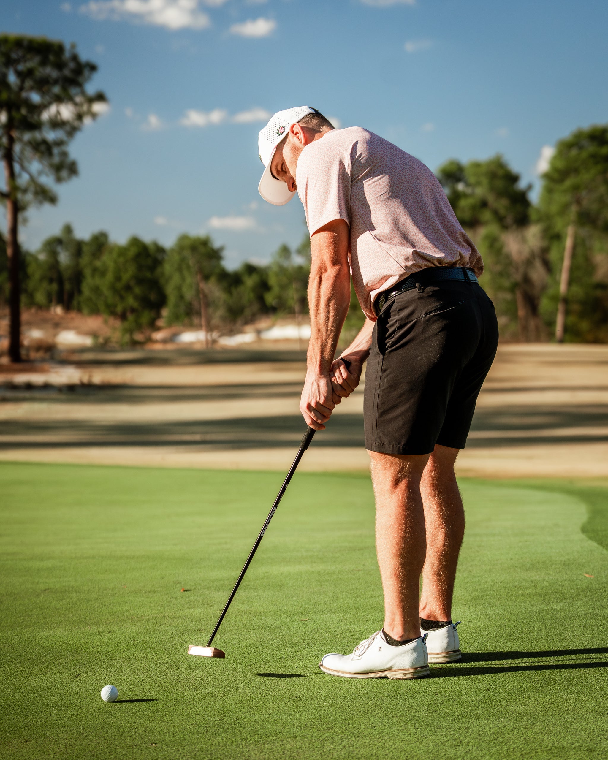 A golfer in a pink polo and black shorts prepares to putt on a sunny course, showcasing stylish golf apparel.