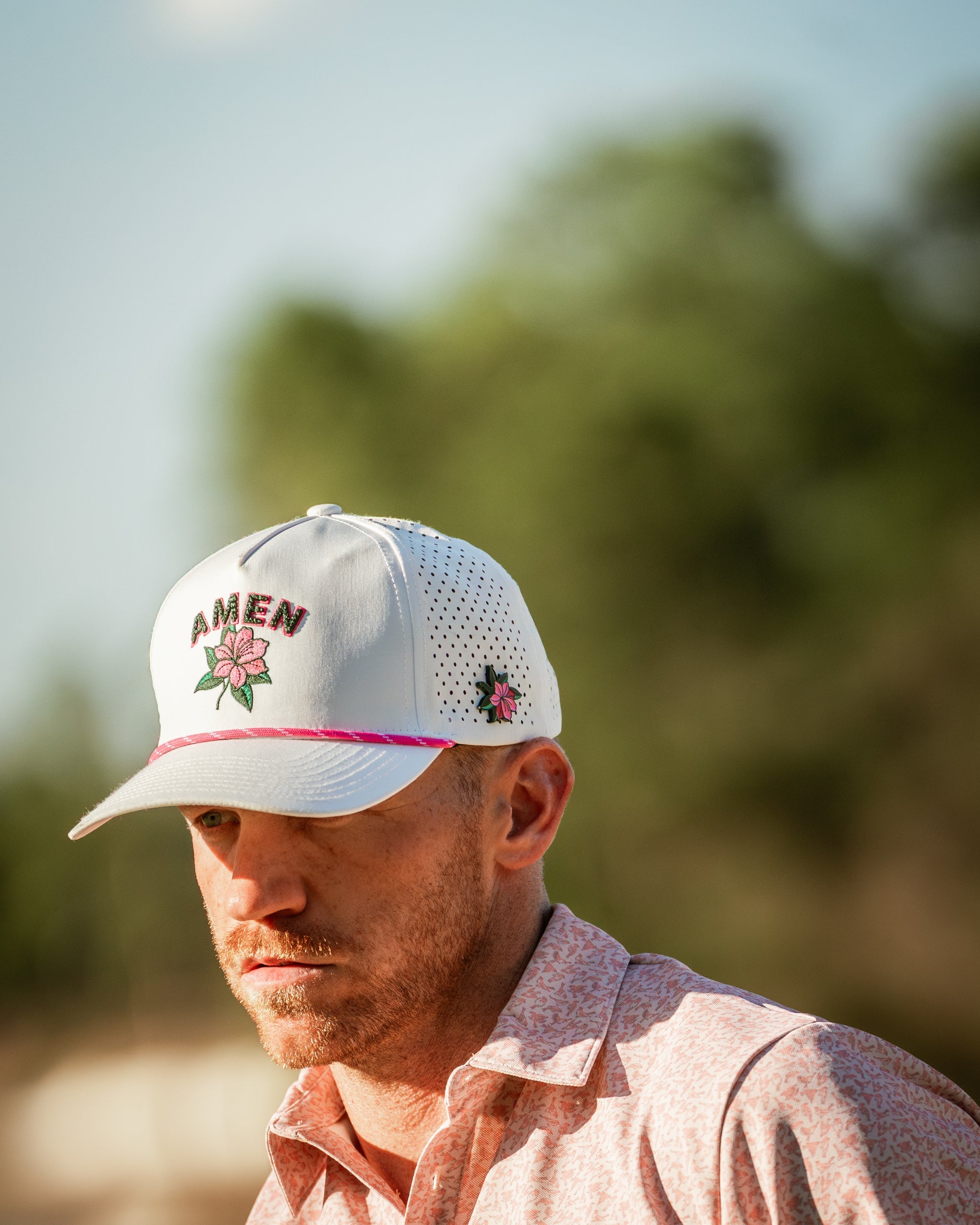 Man wearing a white golf cap with floral embroidery and a pink accent, set against a blurred outdoor background.