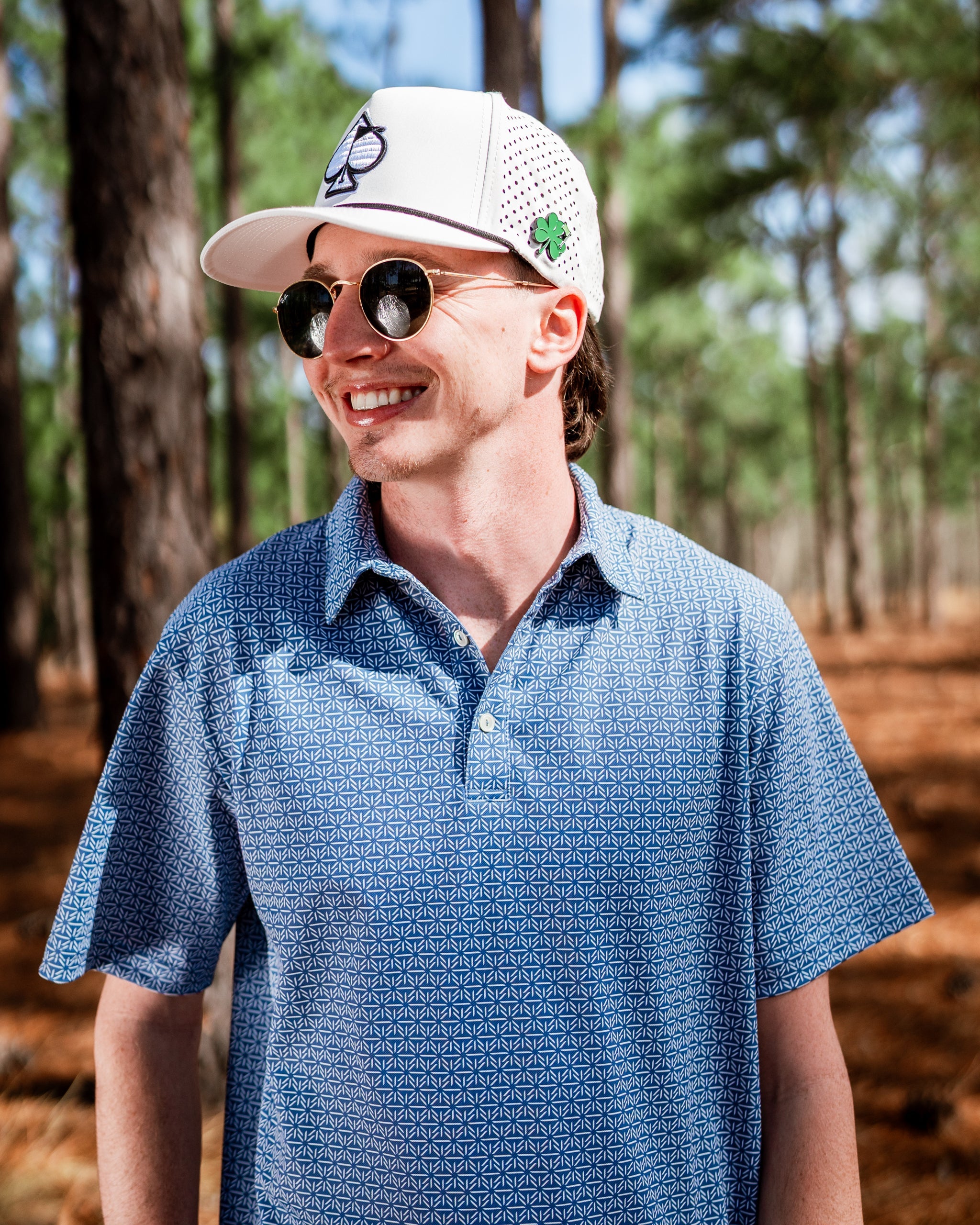 A smiling man wearing a blue patterned polo shirt and a white cap stands among pine trees, showcasing a stylish golf look.