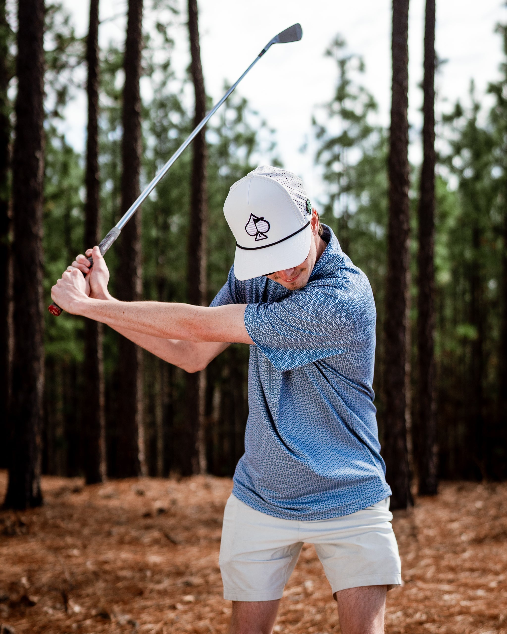 A golfer swings a club in a forested area, wearing a blue patterned polo and a white cap with a unique logo.