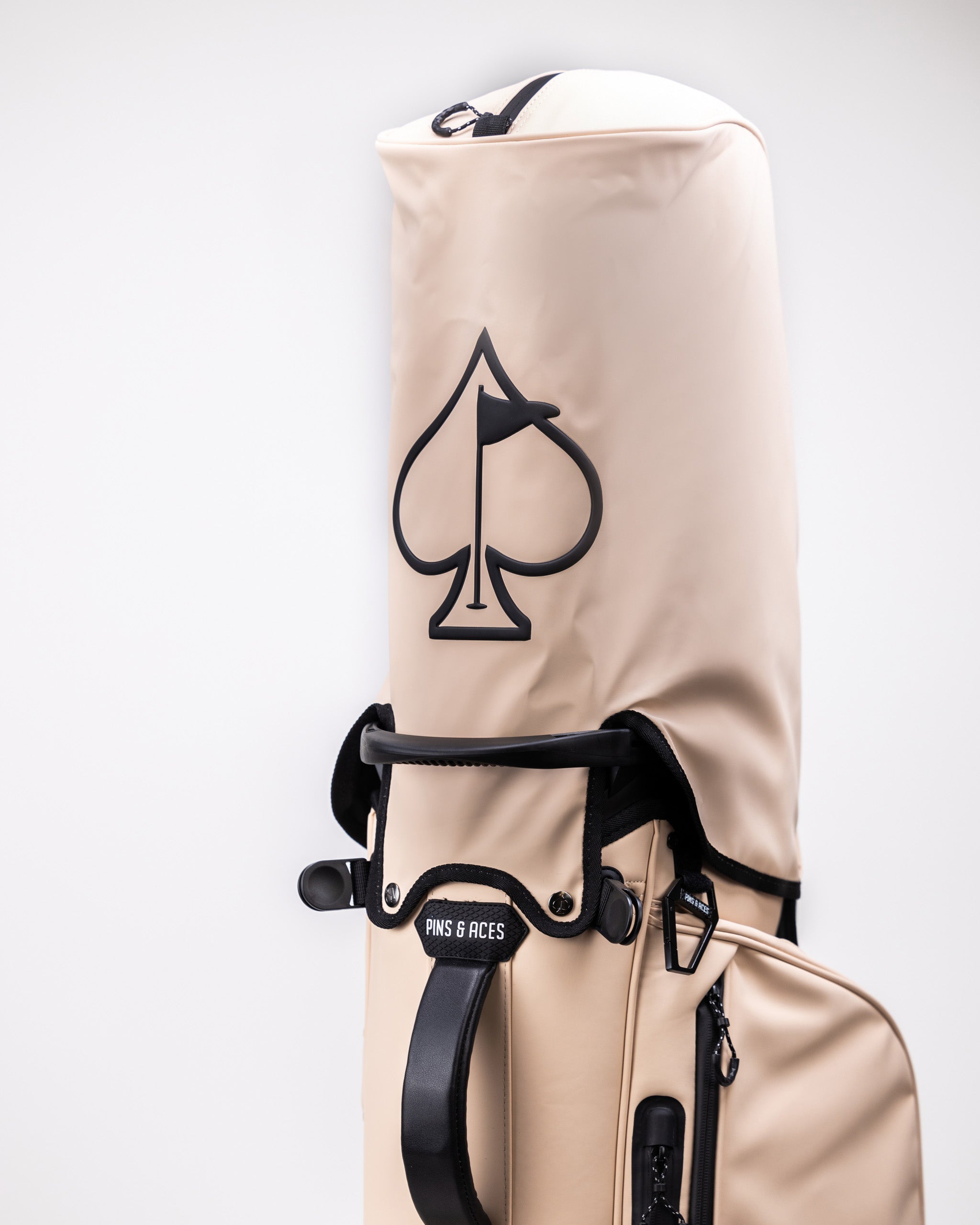 Beige golf bag with black club handle, featuring a spade logo and "Pins & Aces" label for bold, standout golf accessories.