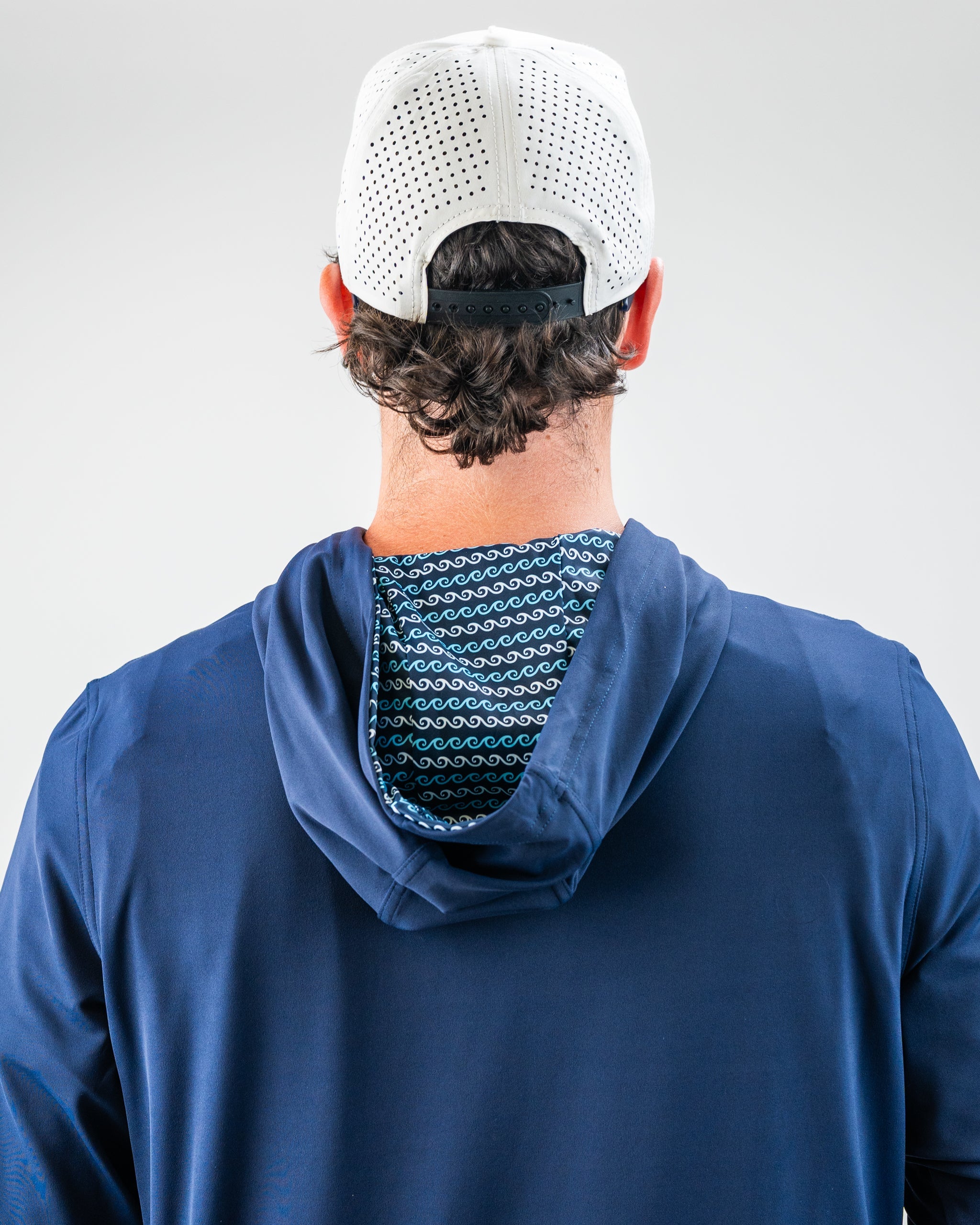 Back view of a man wearing a white perforated golf cap, navy blue jacket, and a patterned hood with blue and white waves.