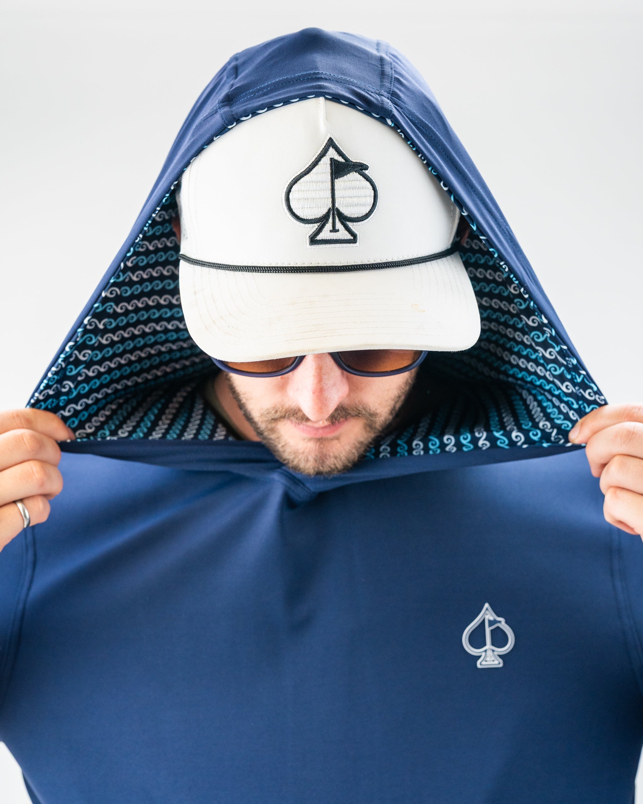 Man wearing a navy blue golf hoodie with a spade logo, lifting the hood, with a white cap featuring a spade and sailboat e...