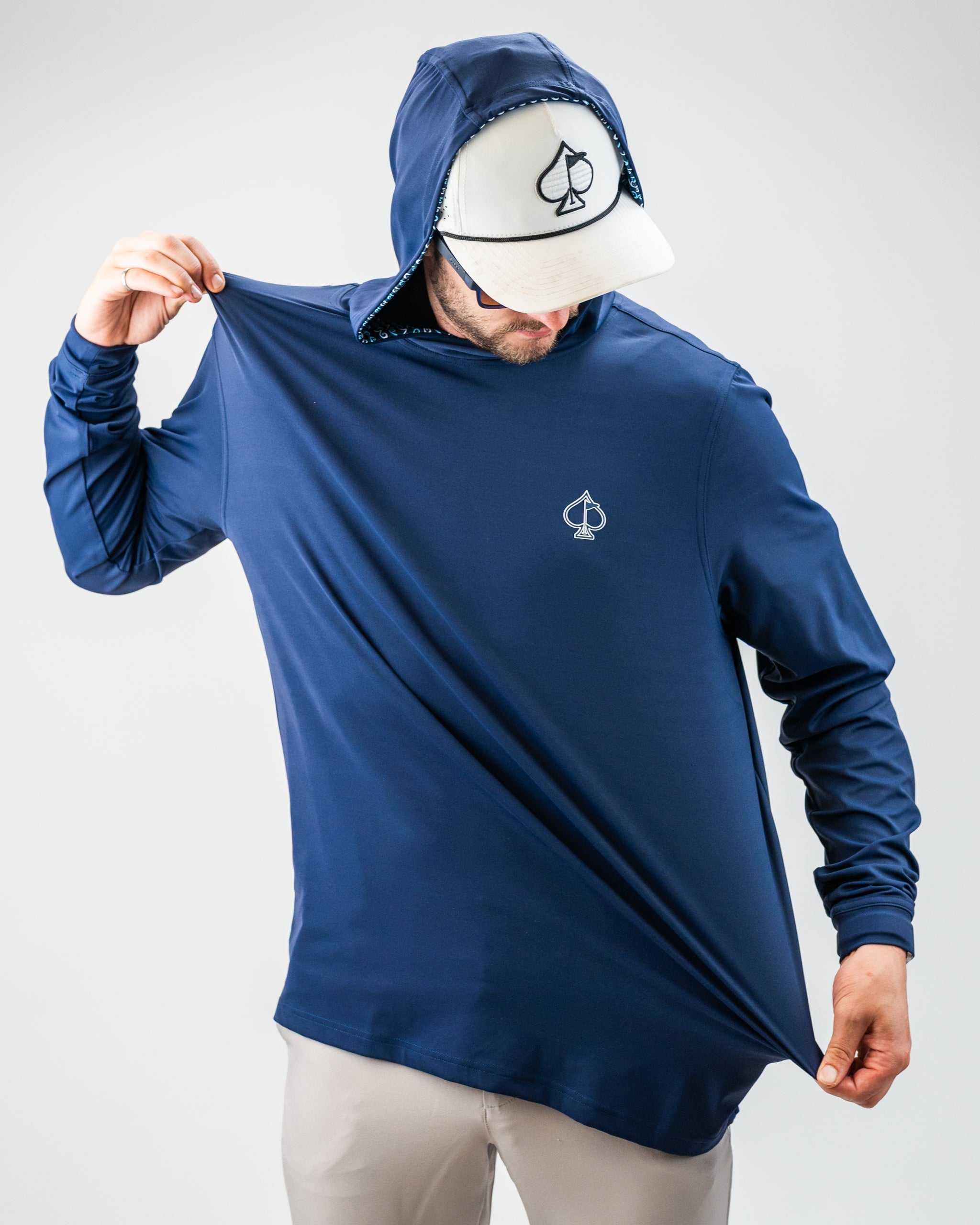 Man wearing a navy blue golf jacket with a spade logo, pulling the fabric to show its stretch, paired with a white cap wit...
