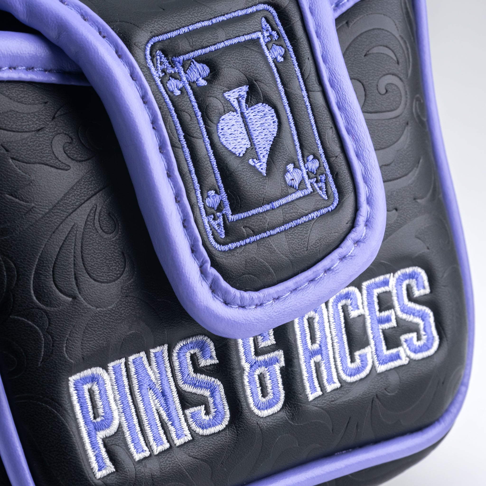 Close-up of a golf mallet putter cover featuring an embroidered Ace of Spades design on a purple and black padded fabric with the text "Pins & Hces" in stylized embroidery.