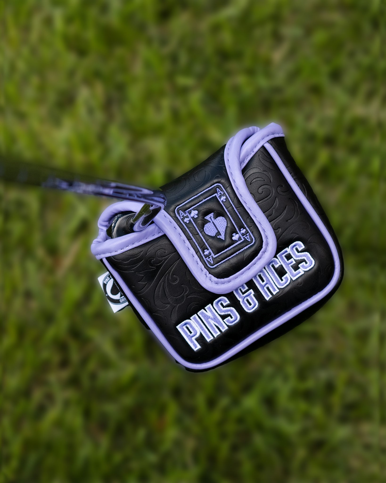 Alt text: "Ace of Spades mallet putter cover by Pins & Aces, featuring a detailed embroidered design on black and purple padded fabric, set against a grassy background."
