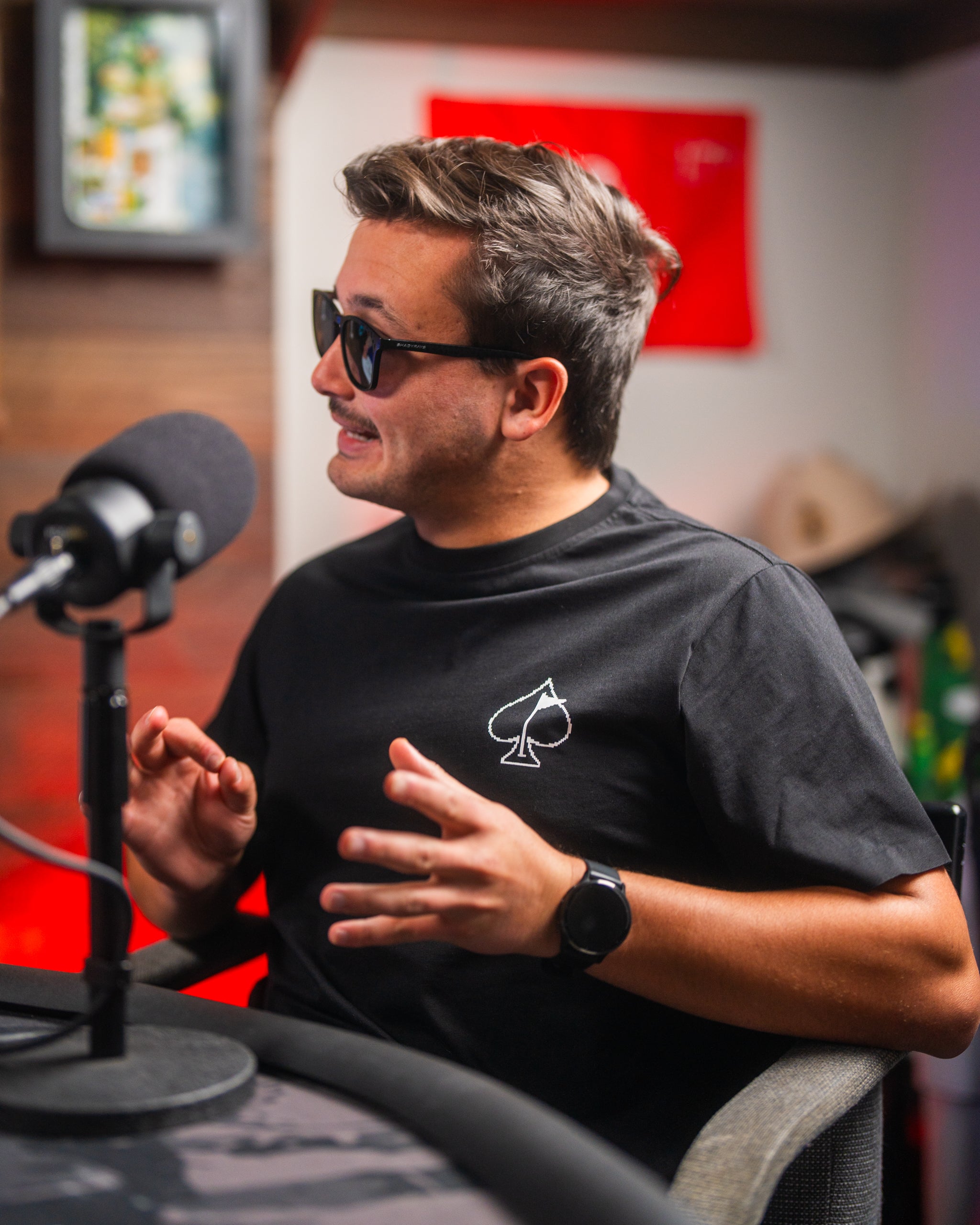 Man wearing a black T-shirt with a white spade logo, sitting in a podcast studio, gesturing while speaking into a microphone, wearing sunglasses and a smartwatch.