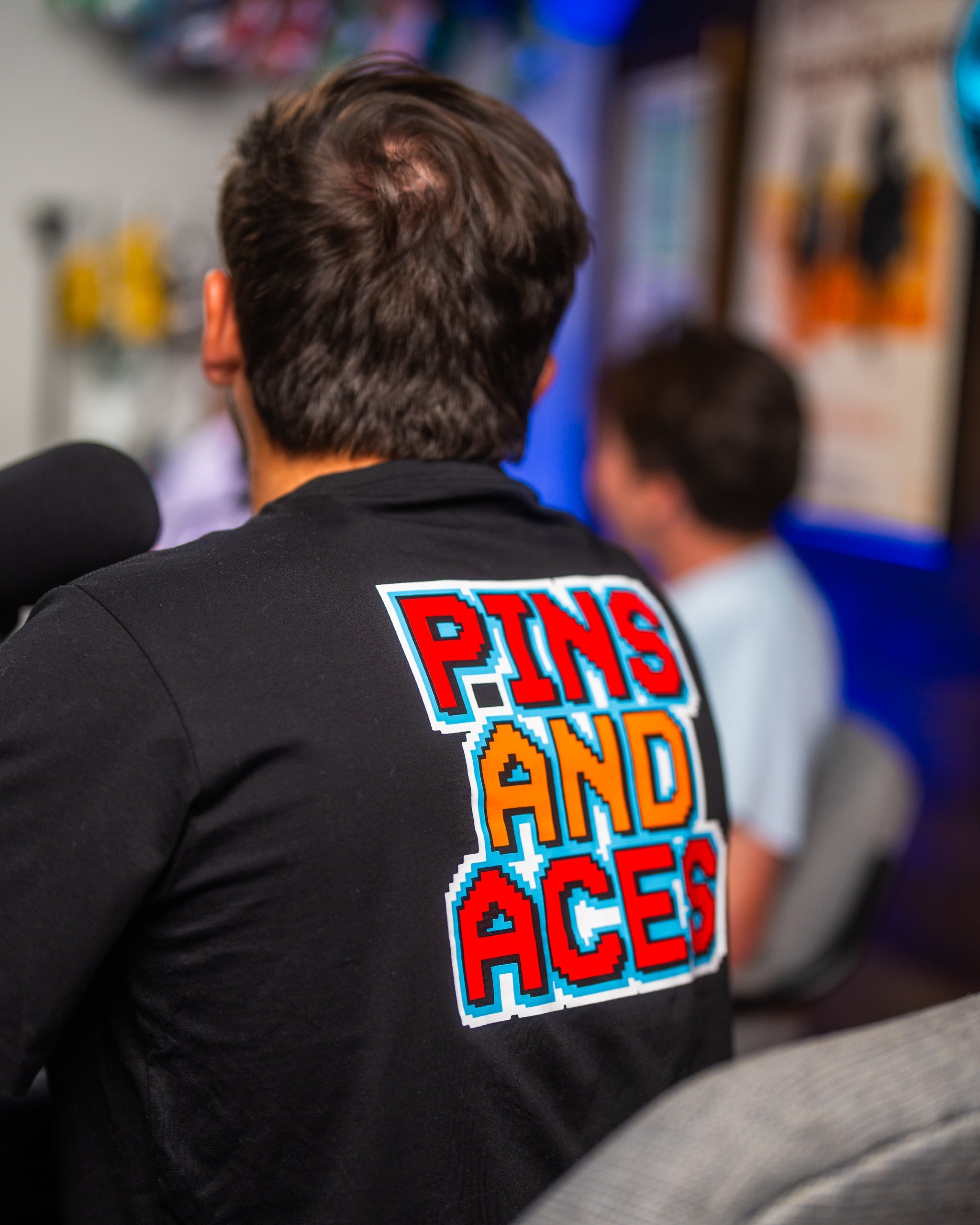 Man wearing a black t-shirt with a colorful "PINS AND ACES" 8-bit retro style graphic on the back, in an arcade setting.