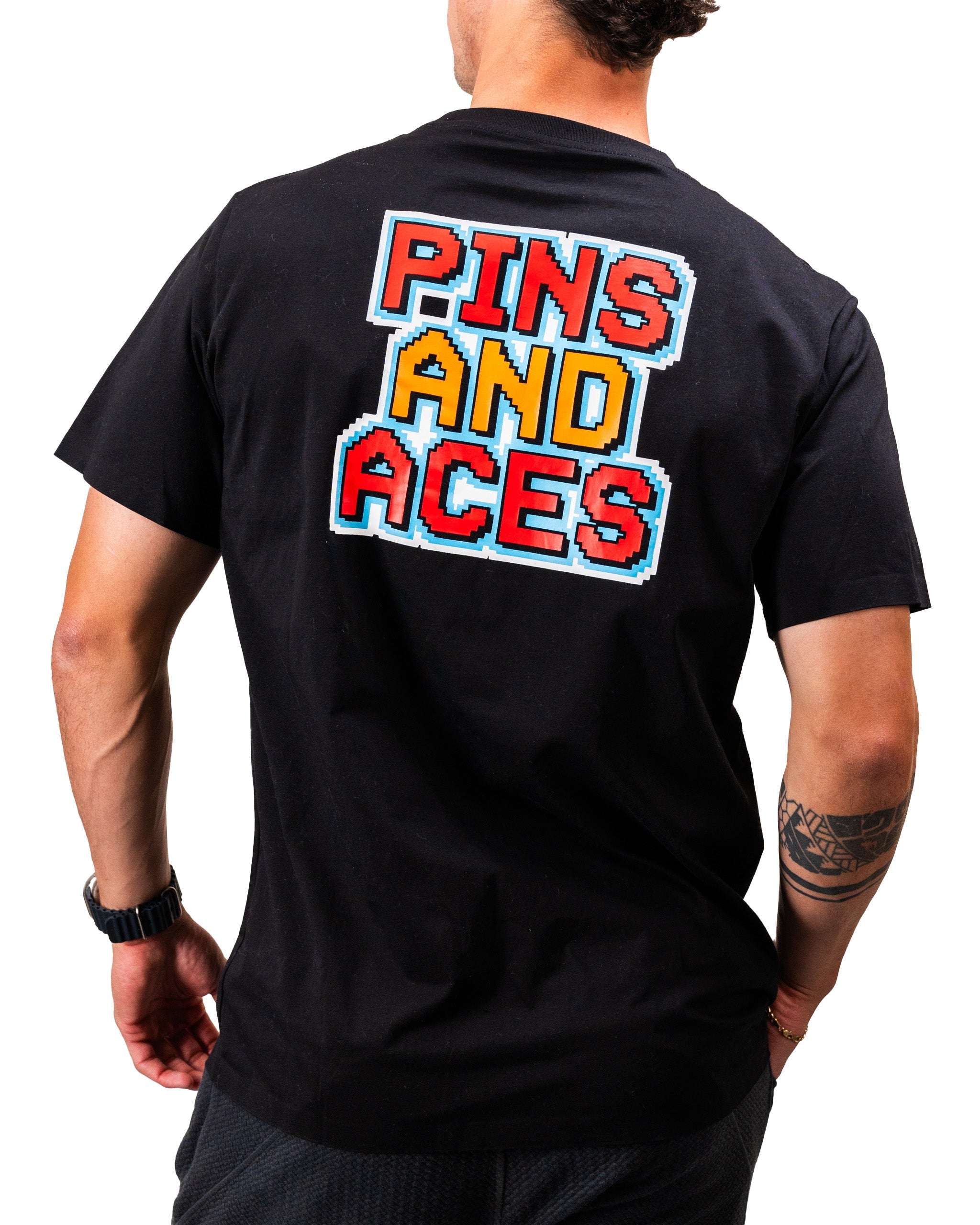 Man wearing a black t-shirt with vibrant 8-bit style graphic text "PINS AND ACES" on the back.