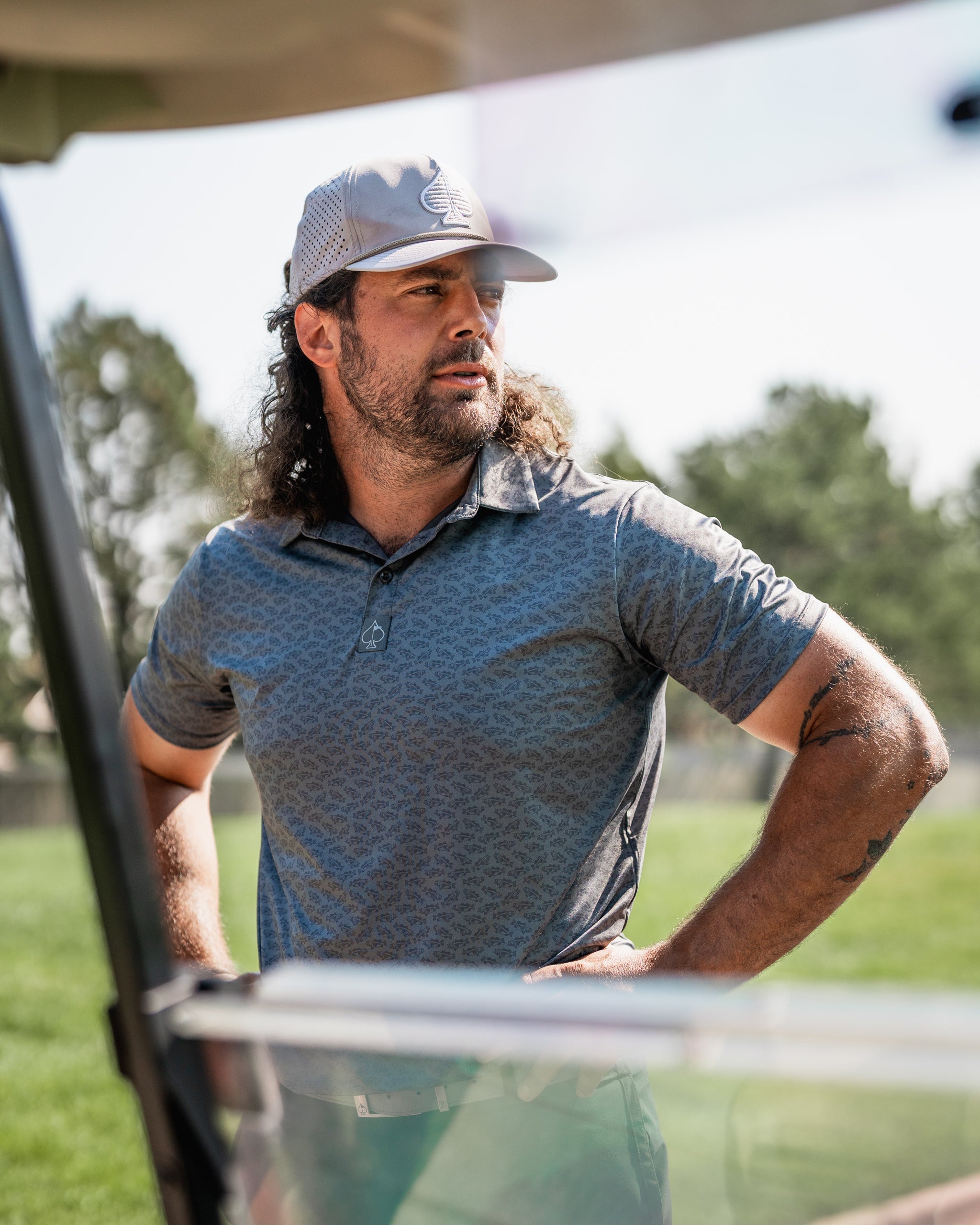 Man in a patterned golf polo shirt stands confidently by a golf cart on a sunny day, ready for his next shot.