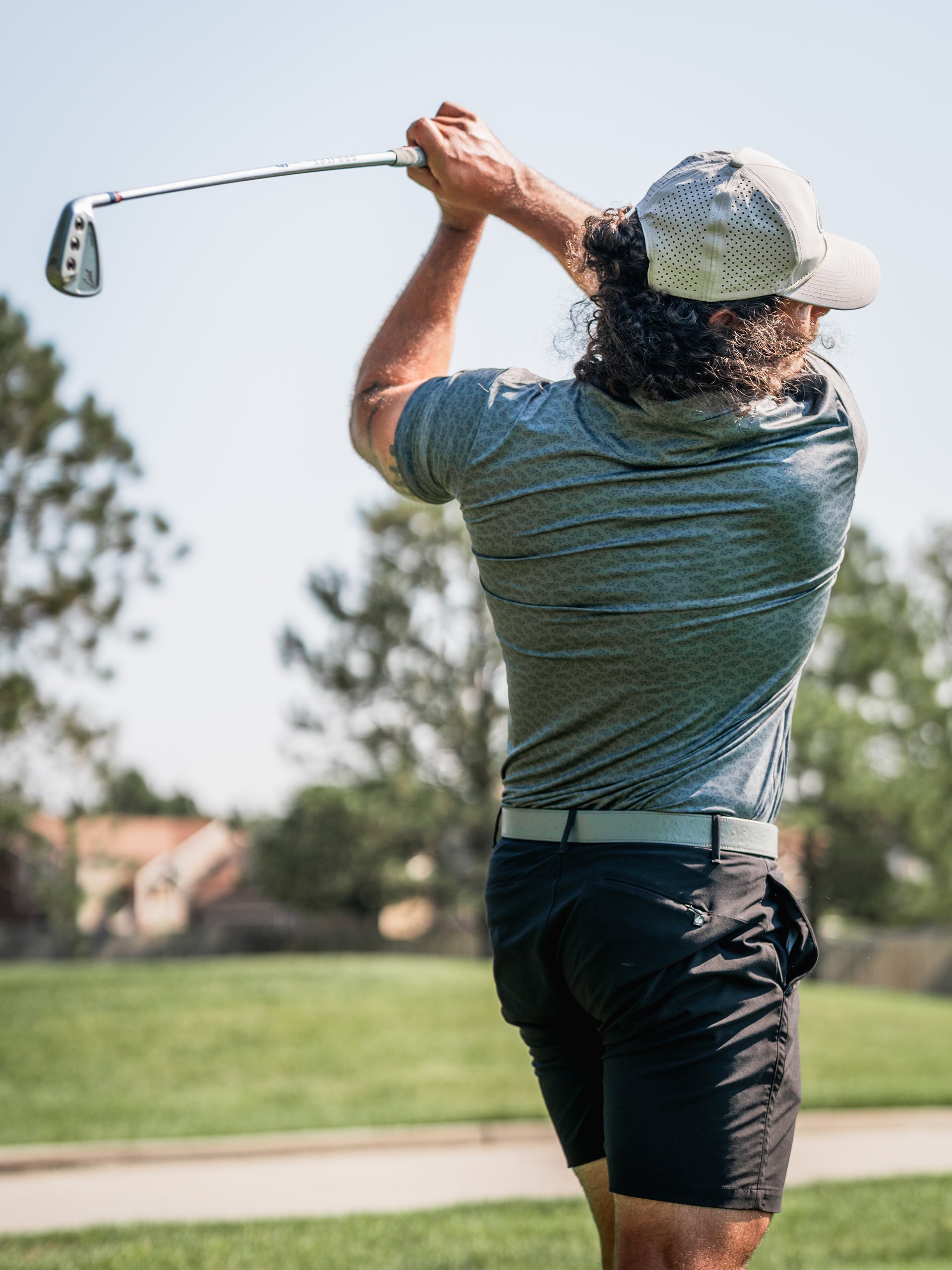 Golfer in a gray shirt and black shorts swings a club on a sunny course, showcasing bold golf apparel in action.