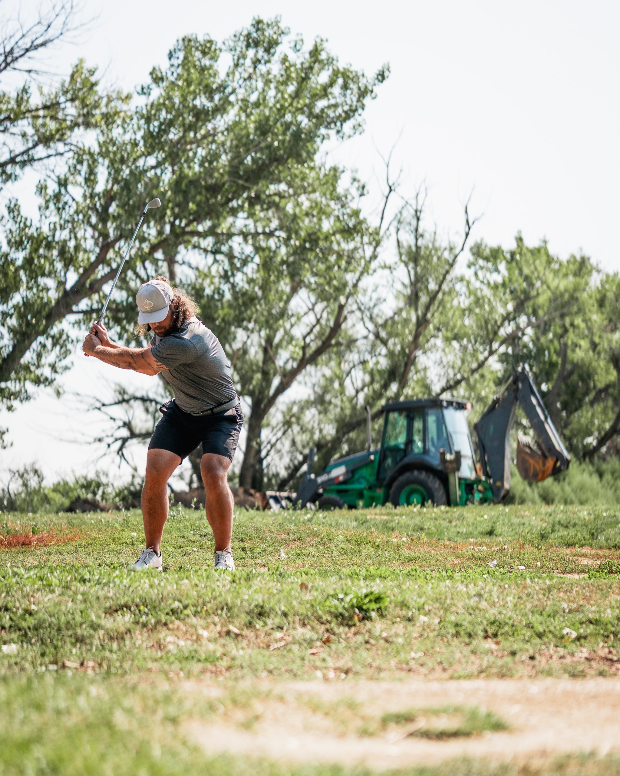 Golfer in a gray shirt and cap swings a club on a grassy course with a tractor in the background.