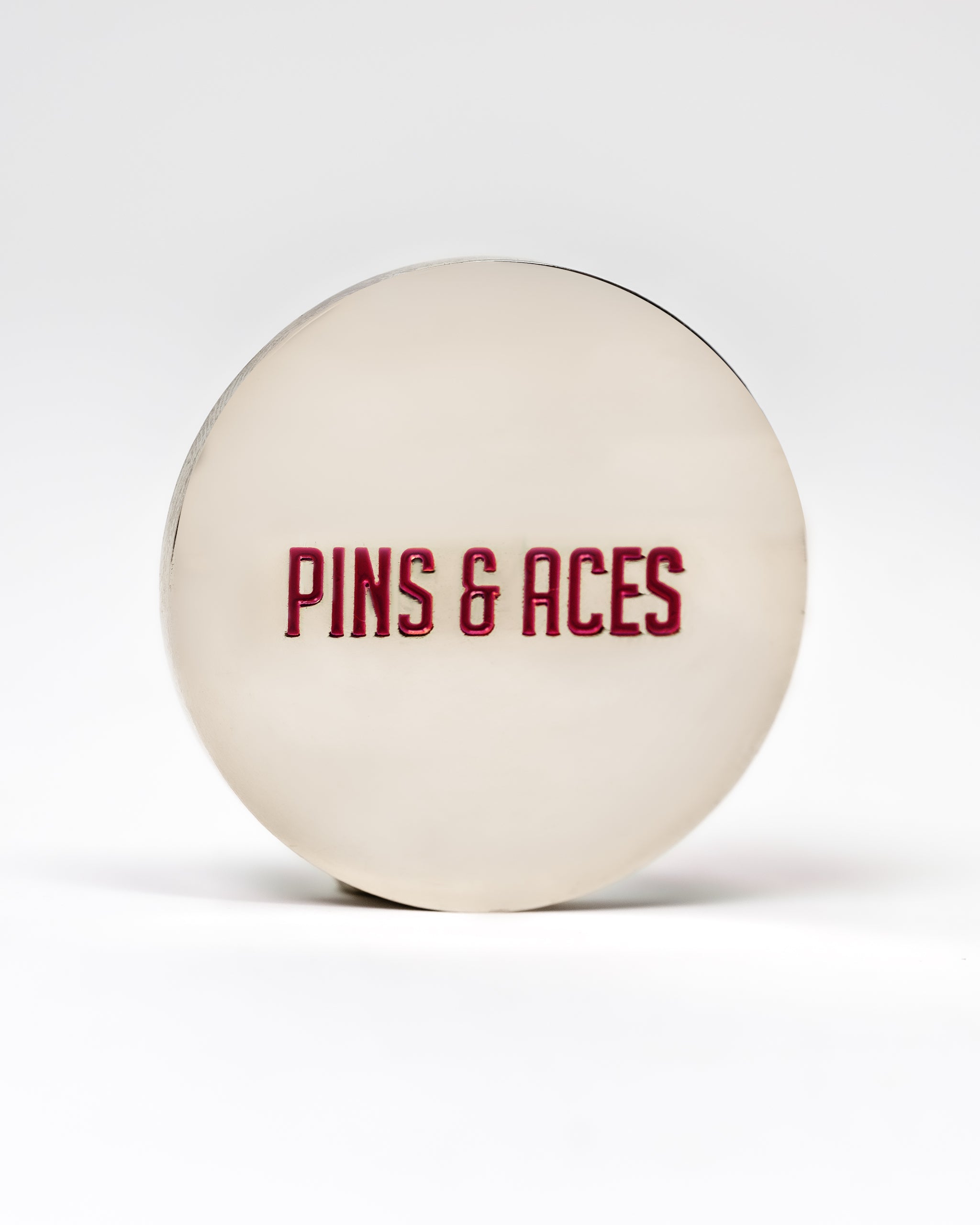 Silver ball marker with "PINS & ACES" embossed in red, designed for golf enthusiasts.