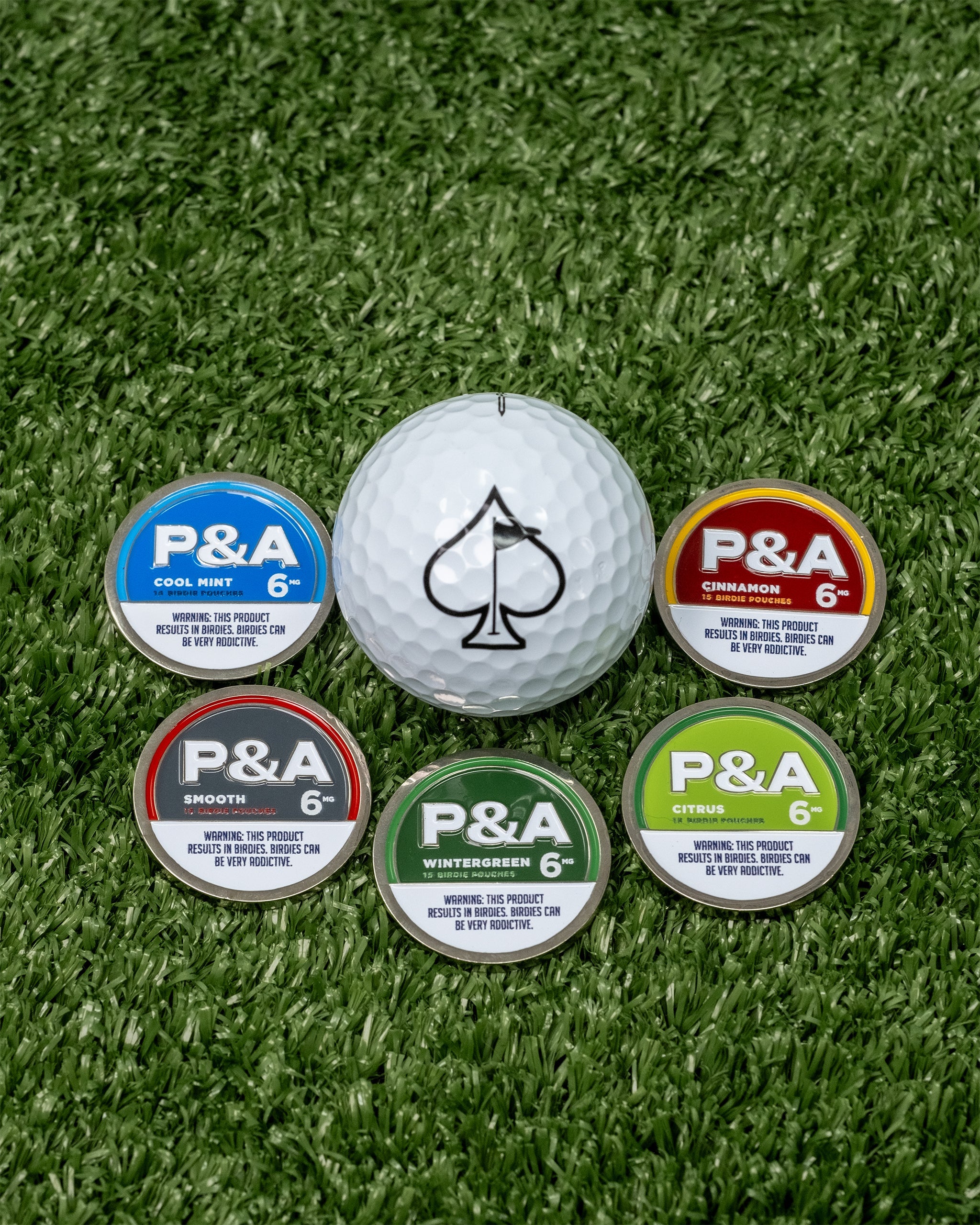 Alt text: "A display of a white golf ball with a spade symbol next to five colorful tins labeled P&A in different flavors: Cool Mint, Cinnamon, Smooth, Wintergreen, and Citrus, all placed on a green grass