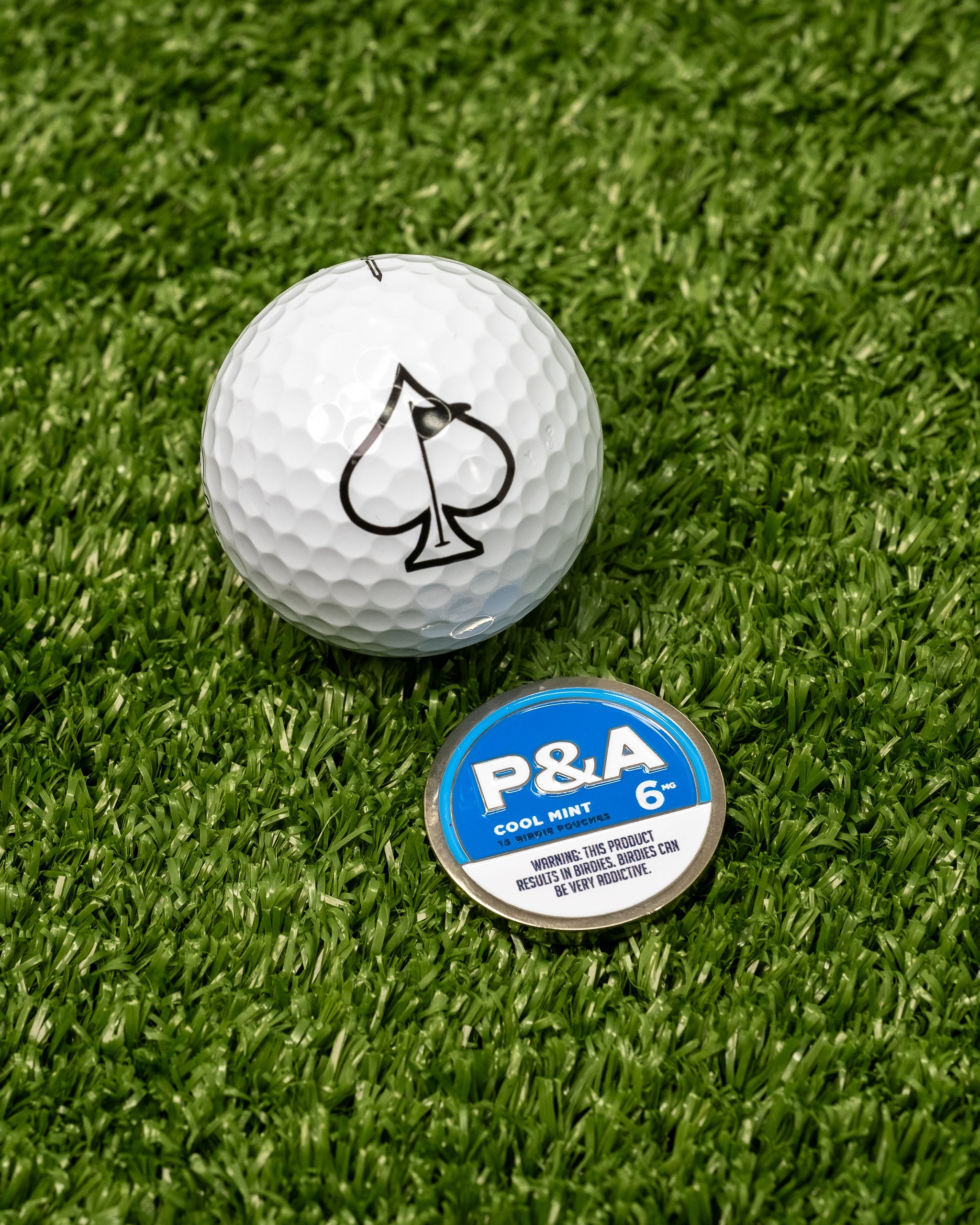 "6 Milly Cool Mint ball marker next to a golf ball with an ace of spades logo on a green turf background.